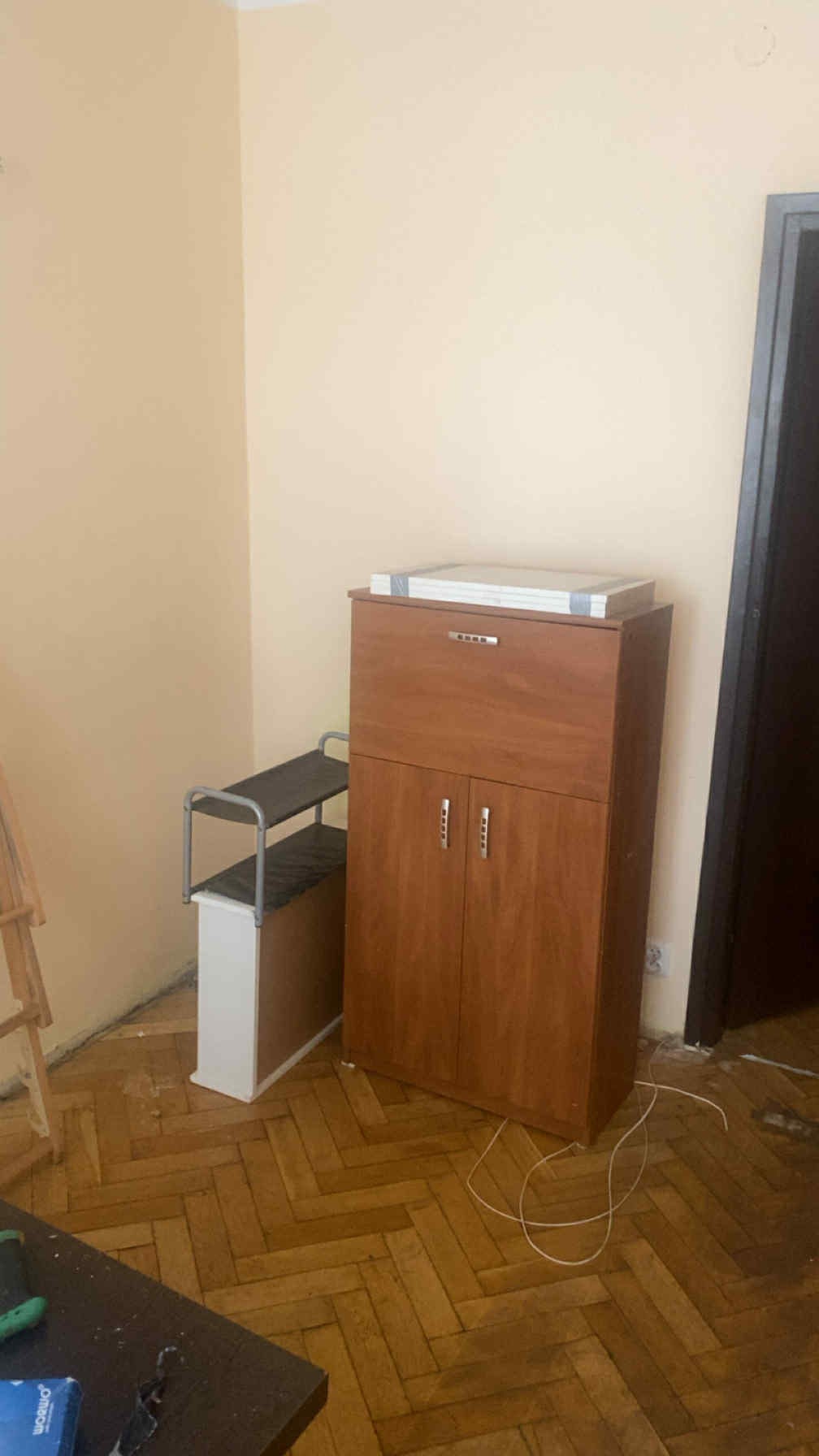 Mebel z ciemnego drewna i metalowy regał na tle beżowej ściany i parkietu. Na szafce leżą zapakowane elementy. Widoczne kable na podłodze.