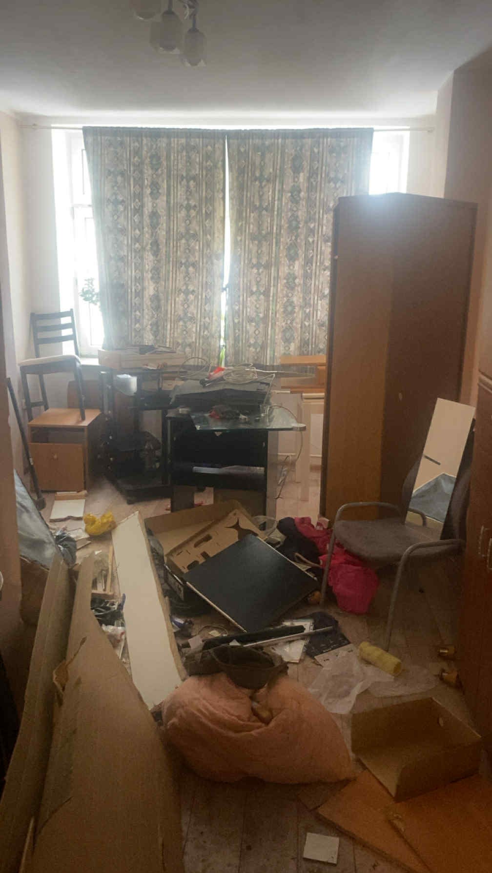 Pokój zagracony kartonami, meblami i różnymi przedmiotami przed opróżnianiem. Widoczne okno z zasłonami i fragmenty innych pomieszczeń. Chaos i bałagan.