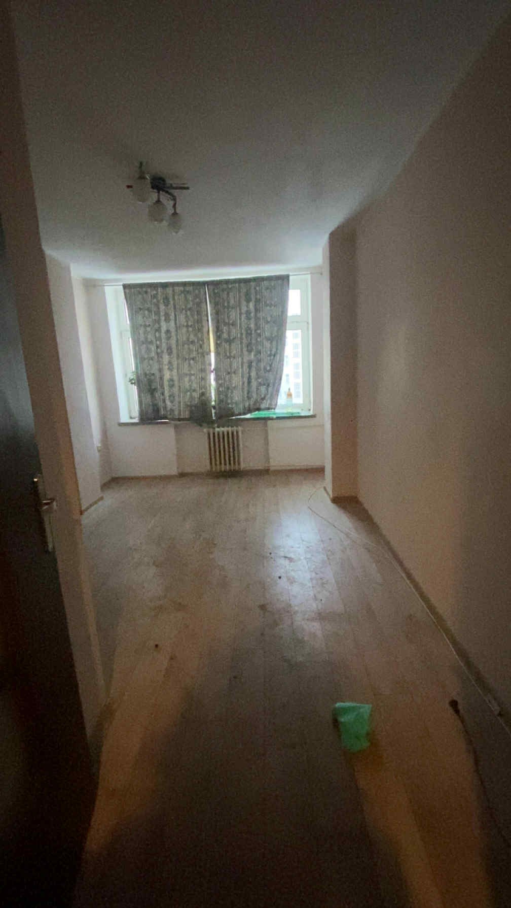 Pusty pokój z drewnianą podłogą, grzejnikiem pod oknem z zasłonami i lampą na suficie. Widoczne ślady użytkowania i pozostałości po remoncie. Neutralne, jasne ściany.