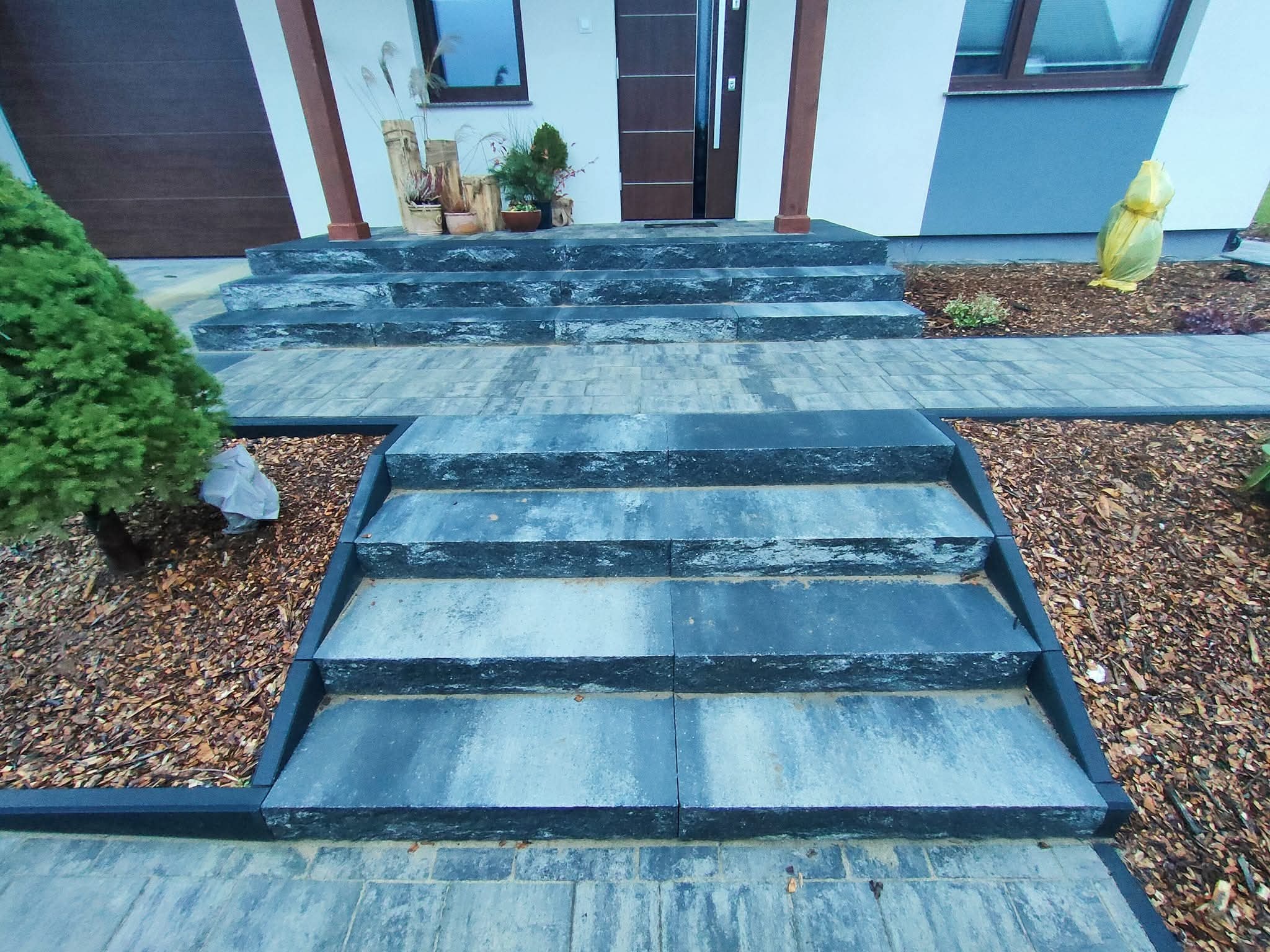 Schody wejściowe z kostki brukowej w odcieniach szarości, z obrzeżami. Tło stanowi elewacja domu z drewnianymi elementami i roślinność.