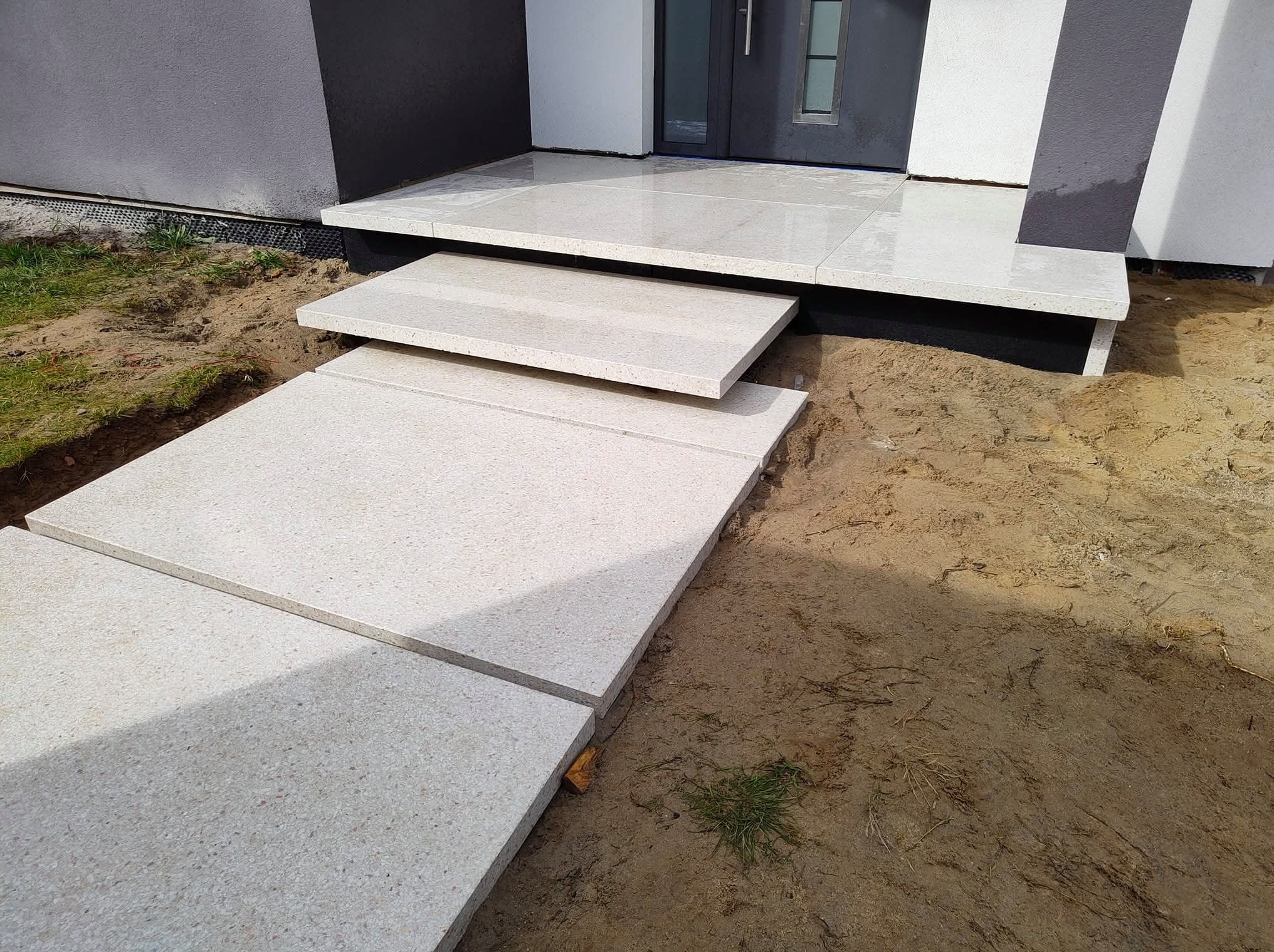 Nowoczesne schody wejściowe z jasnoszarej kostki granitowej prowadzą do drzwi domu, kontrastując z ziemistym otoczeniem. Widoczne detale wykończenia.