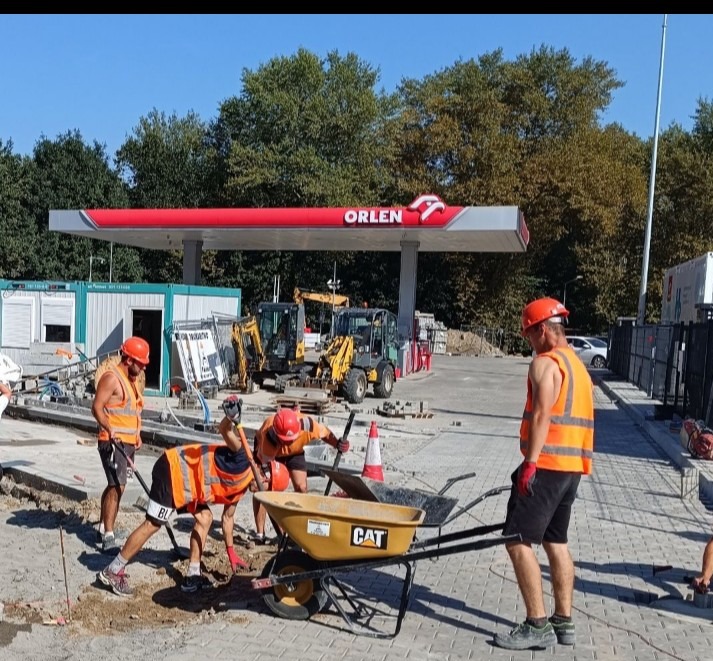 Prace brukarskie na terenie stacji Orlen: robotnicy w kaskach i kamizelkach układają kostkę przy użyciu narzędzi i taczki CAT, z mini koparkami w tle i kontenerami biurowymi.