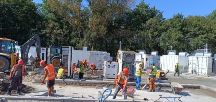 Brygada w pomarańczowych kamizelkach i kaskach pracuje przy układaniu obrzeży na placu budowy. W tle maszyny budowlane i kontenery.