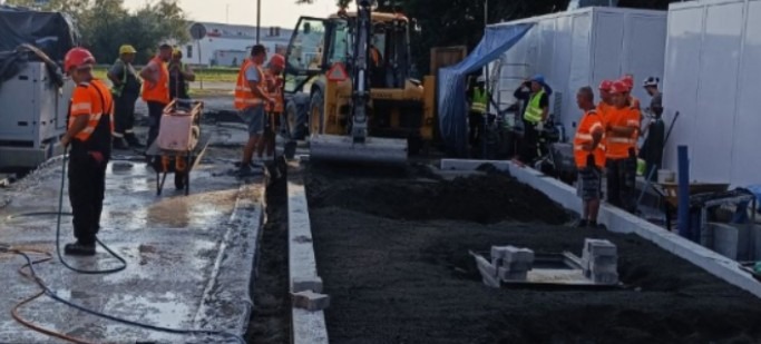 Prace brukarskie w toku: robotnicy w odblaskowych kamizelkach przygotowują podłoże pod kostkę, używając walca i ubijarki. Widoczny etap wyrównywania terenu podłoża.