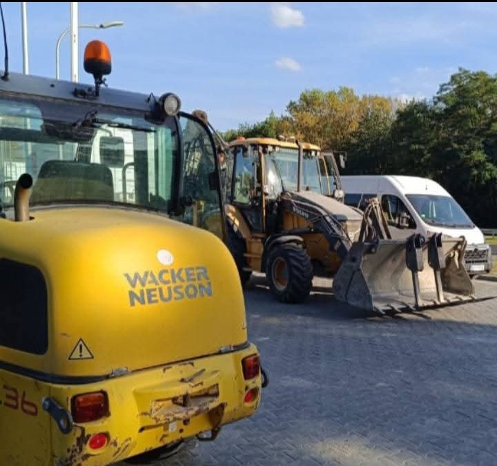 Żółty walec Wacker Neuson, koparko-ładowarka Volvo i biały bus na placu budowy. Sprzęt gotowy do układania kostki brukowej.