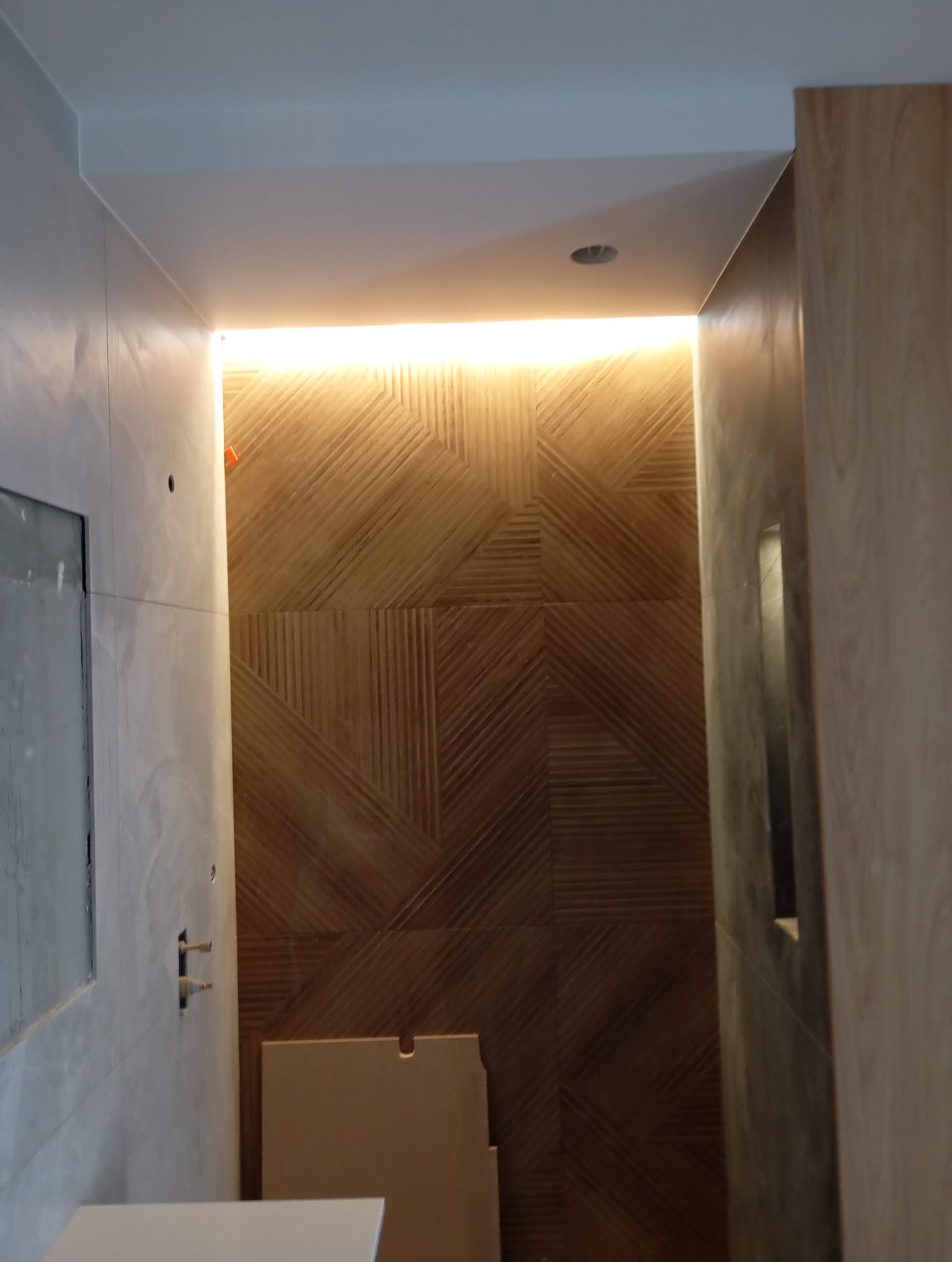 Nowoczesna łazienka z podświetlaną ścianą z drewnianą mozaiką, widok na fragment umywalki i przygotowane podejścia wodne, minimalistyczny design.