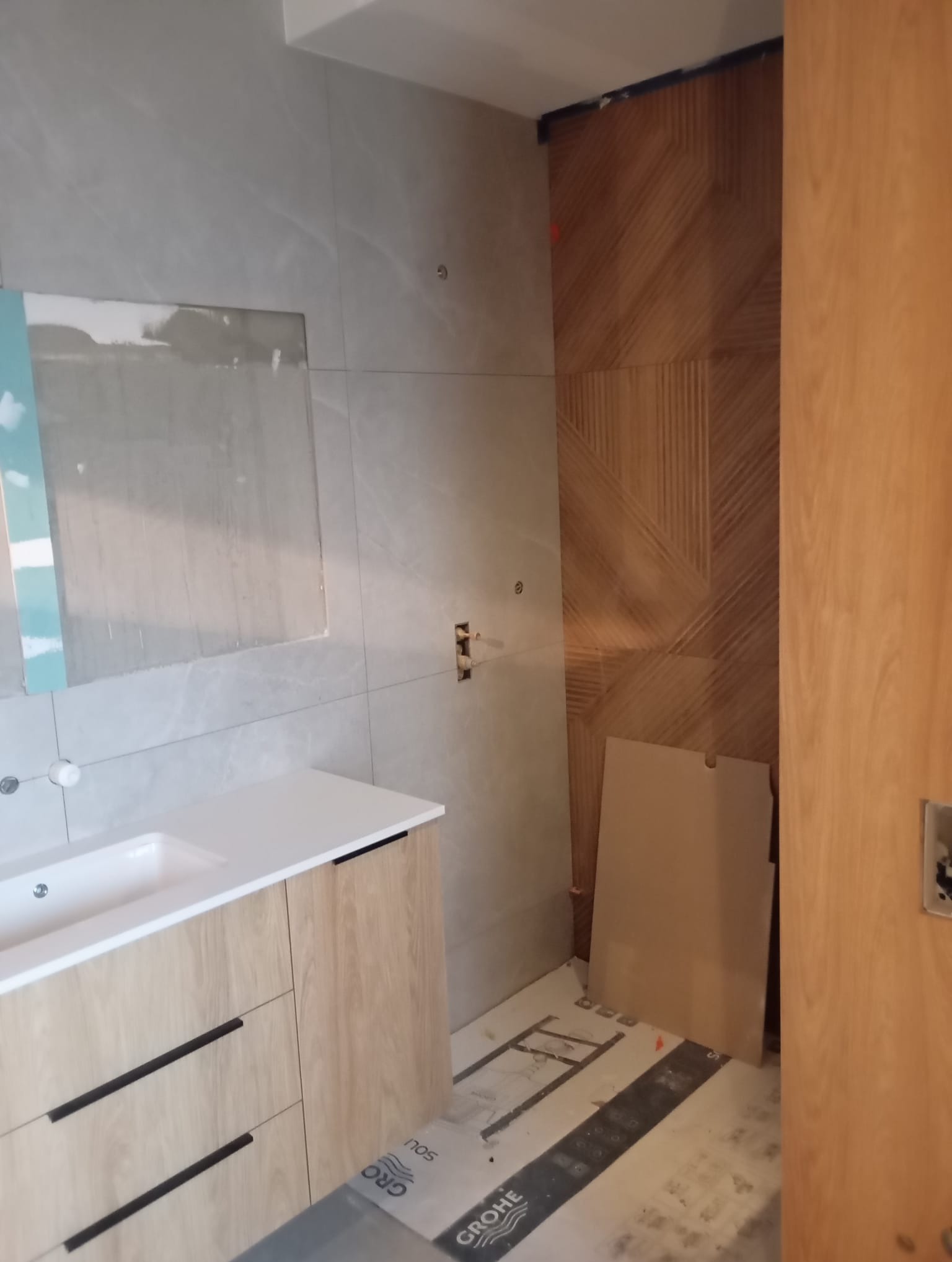 Łazienka w trakcie wykańczania: umywalka z szafką w odcieniach drewna, szare płytki na ścianach i dekoracyjna ściana z drewnianych paneli. Widoczne materiały budowlane.