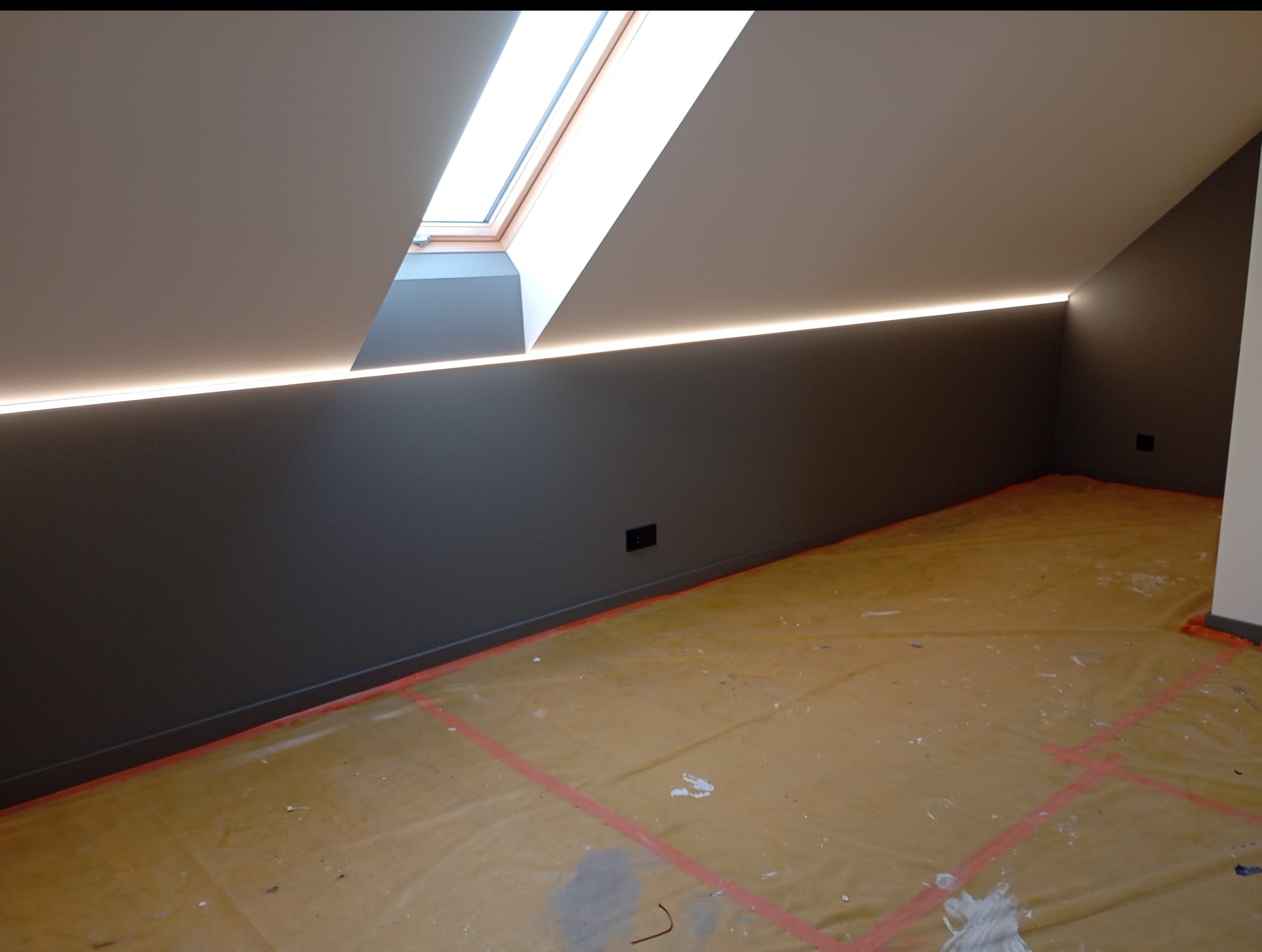 Szara ściana z oknem dachowym i subtelnym oświetleniem LED wzdłuż krawędzi sufitu. Podłoga zabezpieczona folią malarską. Widok na ukończone wykończenie poddasza.