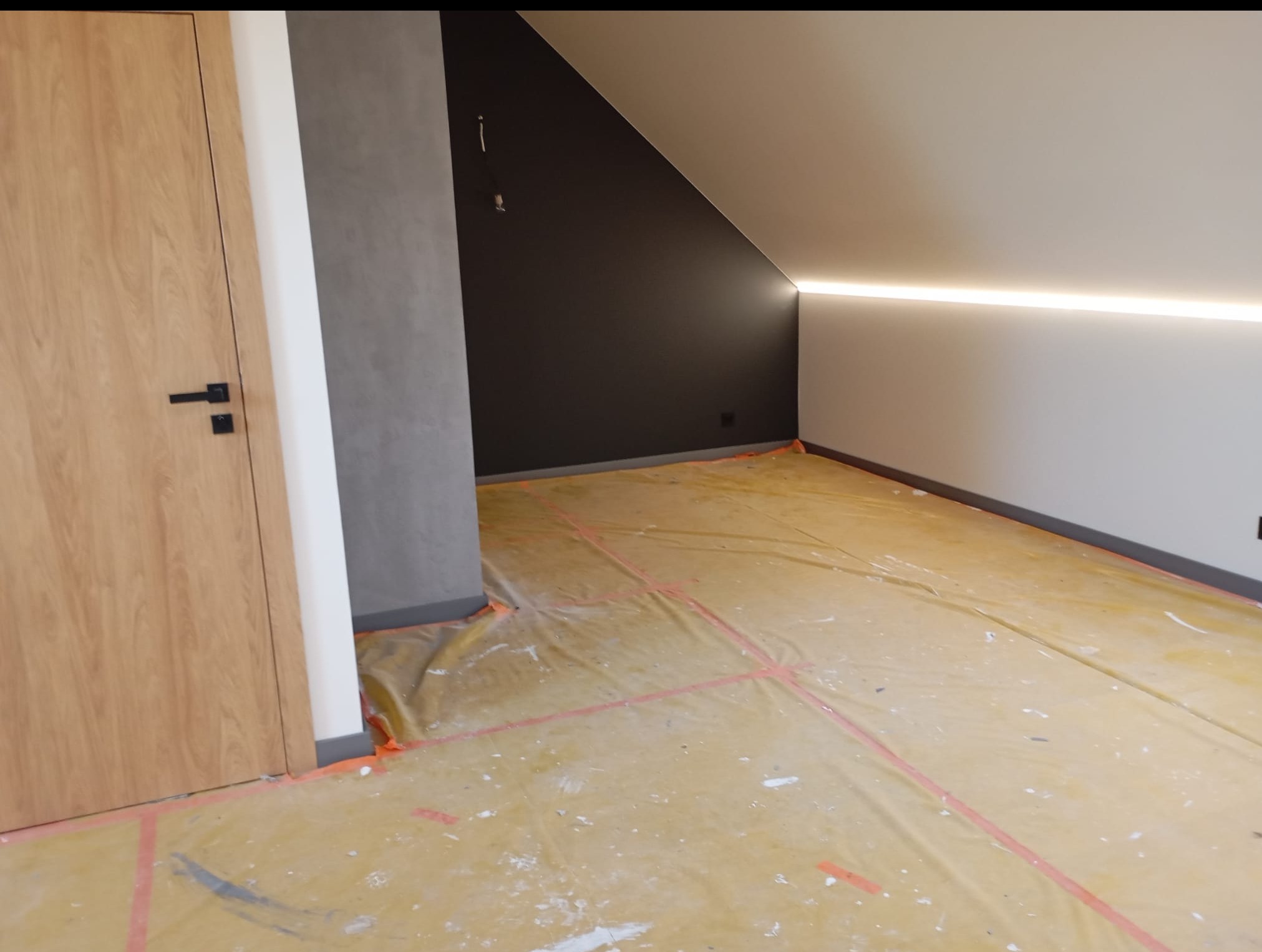 Pokój na poddaszu po malowaniu ścian w odcieniach szarości i bieli, z zamontowaną listwą LED. Podłoga zabezpieczona folią malarską. Widoczna drewniana ościeżnica drzwi.