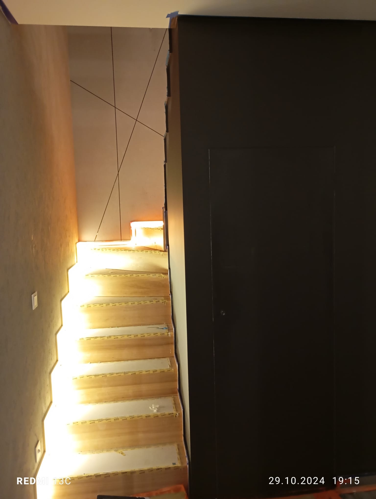 Oświetlone schody z drewnianymi stopniami w trakcie montażu, widoczne taśmy zabezpieczające, minimalistyczny design ściany z ciemnymi liniami, nowoczesne wykończenie wnętrza.