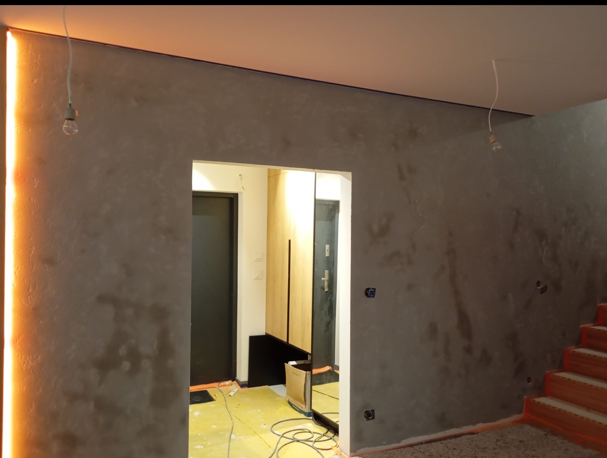 Wnętrze w trakcie wykańczania: szara ściana z efektem dekoracyjnym, surowe żarówki, fragment schodów i widok na przedpokój z lustrem. Podłoga zabezpieczona folią.