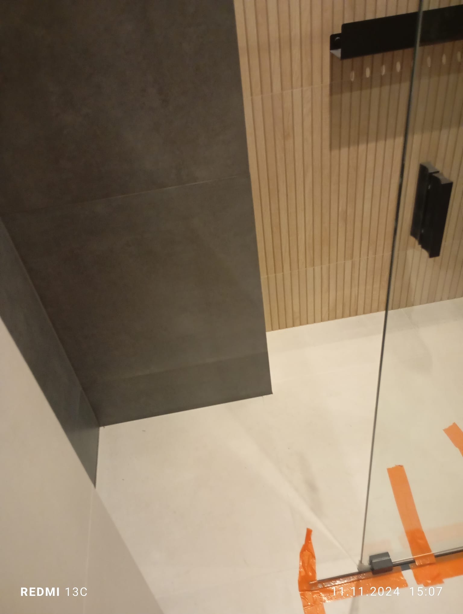 Wykończona łazienka z szarą płytką na suficie i drewnopodobną na ścianie. Widoczne zabezpieczenia folią na podłodze i szklana kabina prysznicowa.