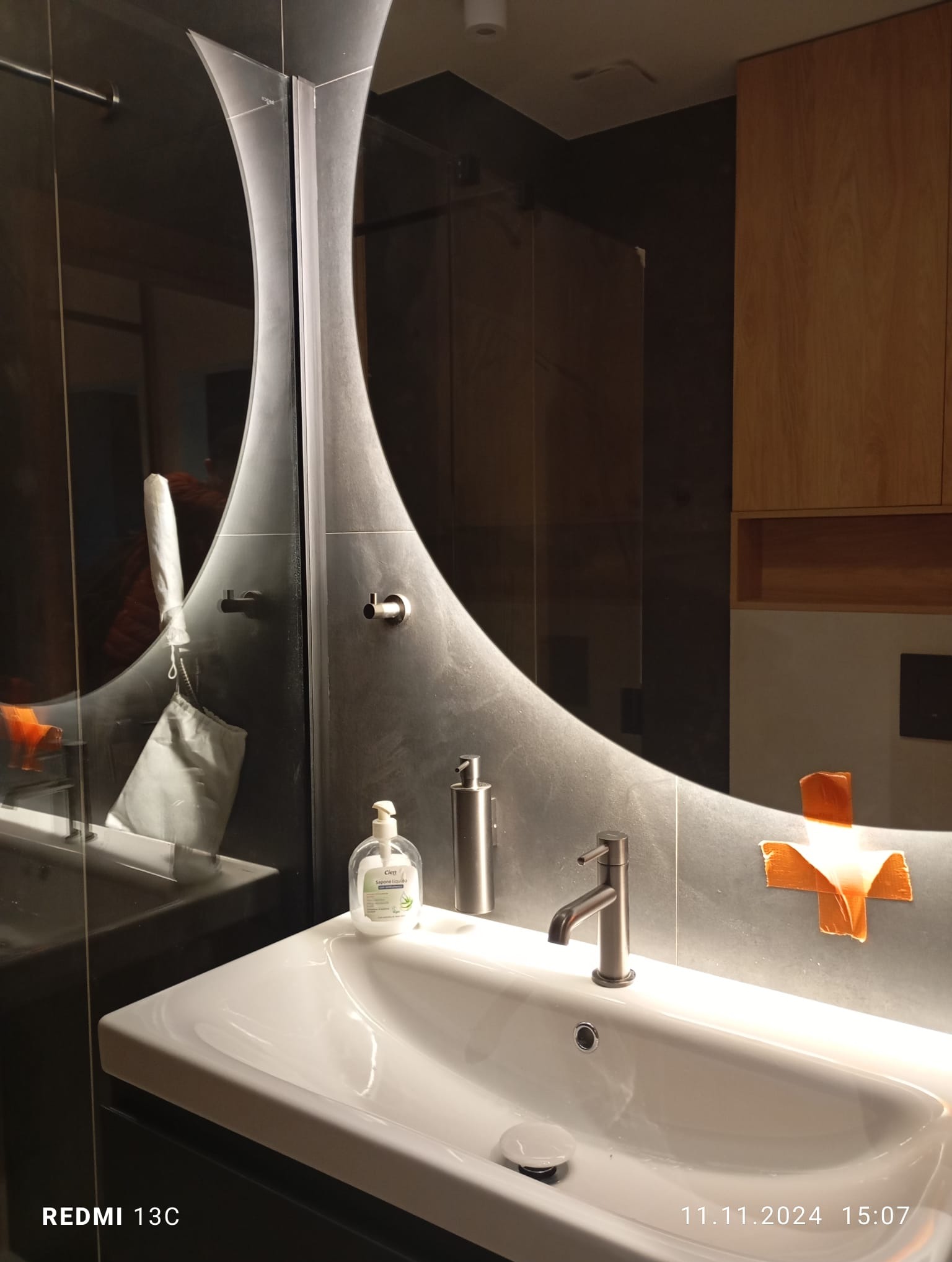 Nowoczesna łazienka z umywalką i armaturą w odcieniach szarości, oświetlona ciepłym światłem. Na blacie dozownik mydła i płynu. W lustrze odbicie kabiny prysznicowej.