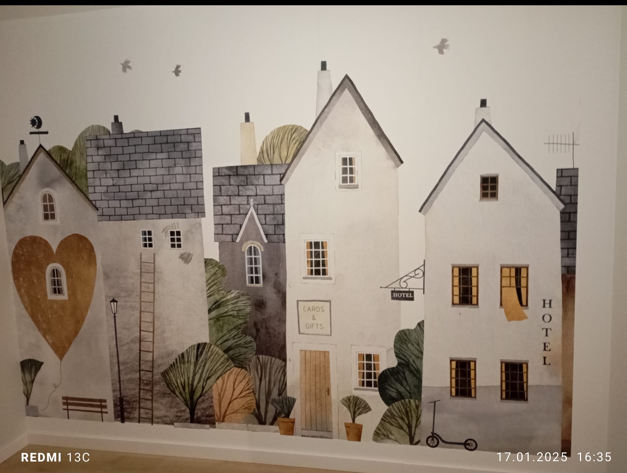 Ściana z dekoracyjnym malowidłem przedstawiającym budynki w stylu akwareli, z subtelnymi detalami jak drabina i rośliny w doniczkach. U dołu widoczna hulajnoga.