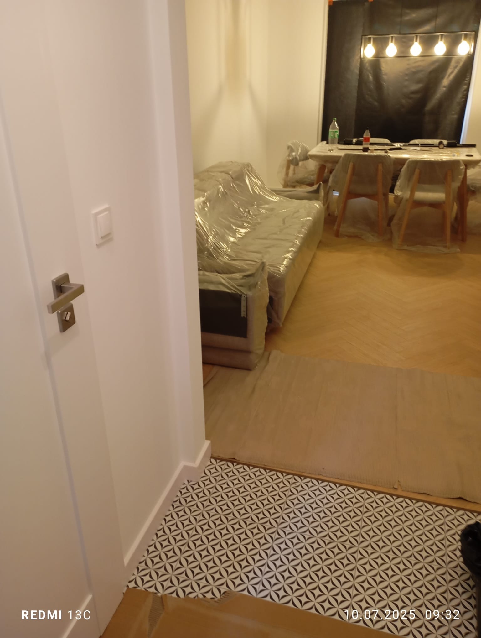Wnętrze w trakcie zabezpieczania folią malarską: sofa, stół i krzesła przykryte, podłoga częściowo zabezpieczona, widoczny fragment nowej podłogi z wzorem.
