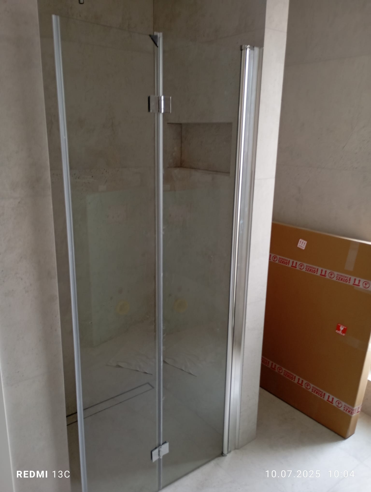 Szklana kabina prysznicowa z chromowanymi okuciami, widoczna na tle wykończonej, jasnej łazienki z pudełkiem po szkle w rogu. Minimalistyczny design.