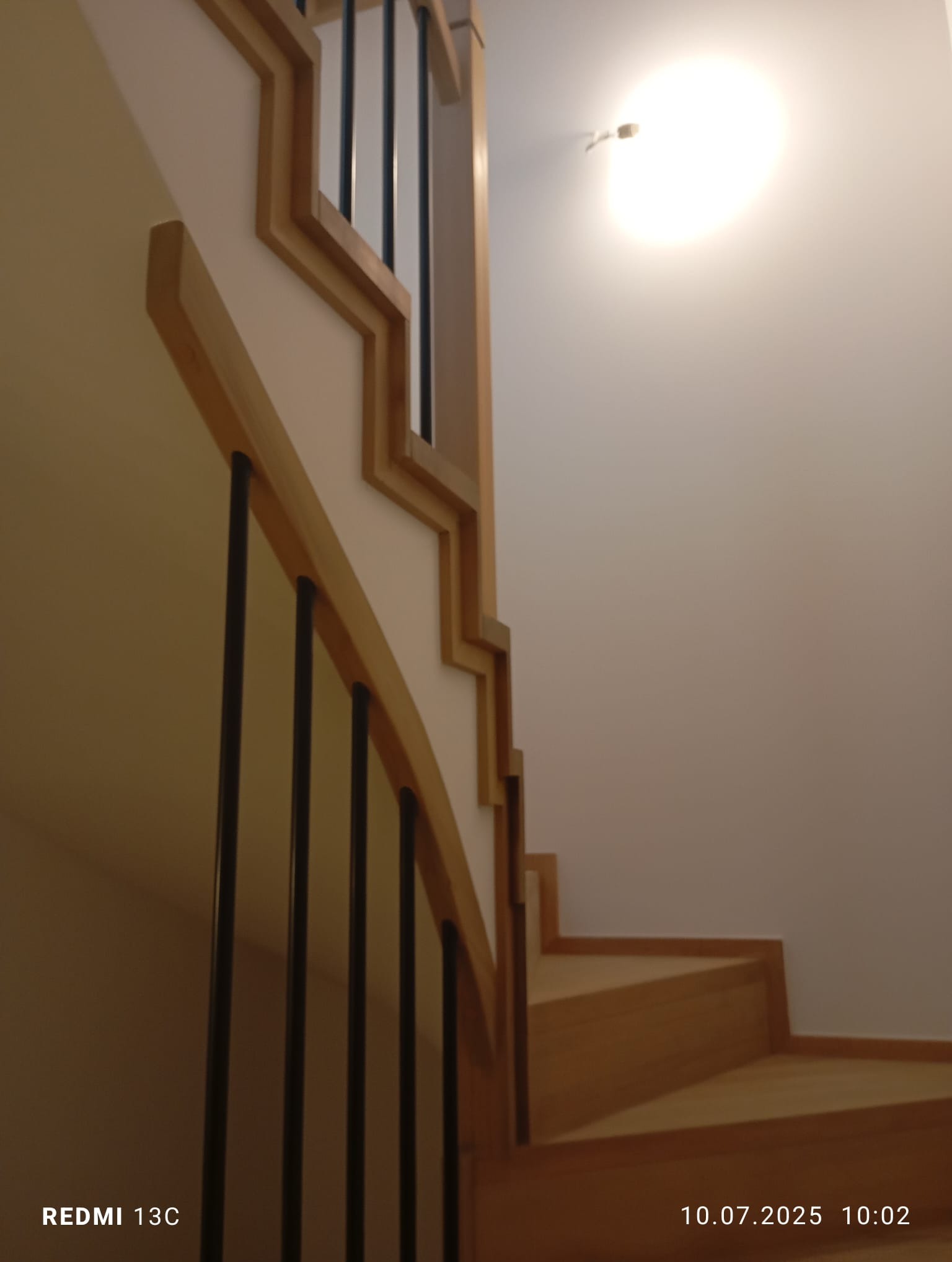 Nowoczesne schody drewniane z czarnymi, metalowymi balustradami i jasnym oświetleniem, widoczne z dołu, ukazujące estetyczne wykończenie wnętrza.