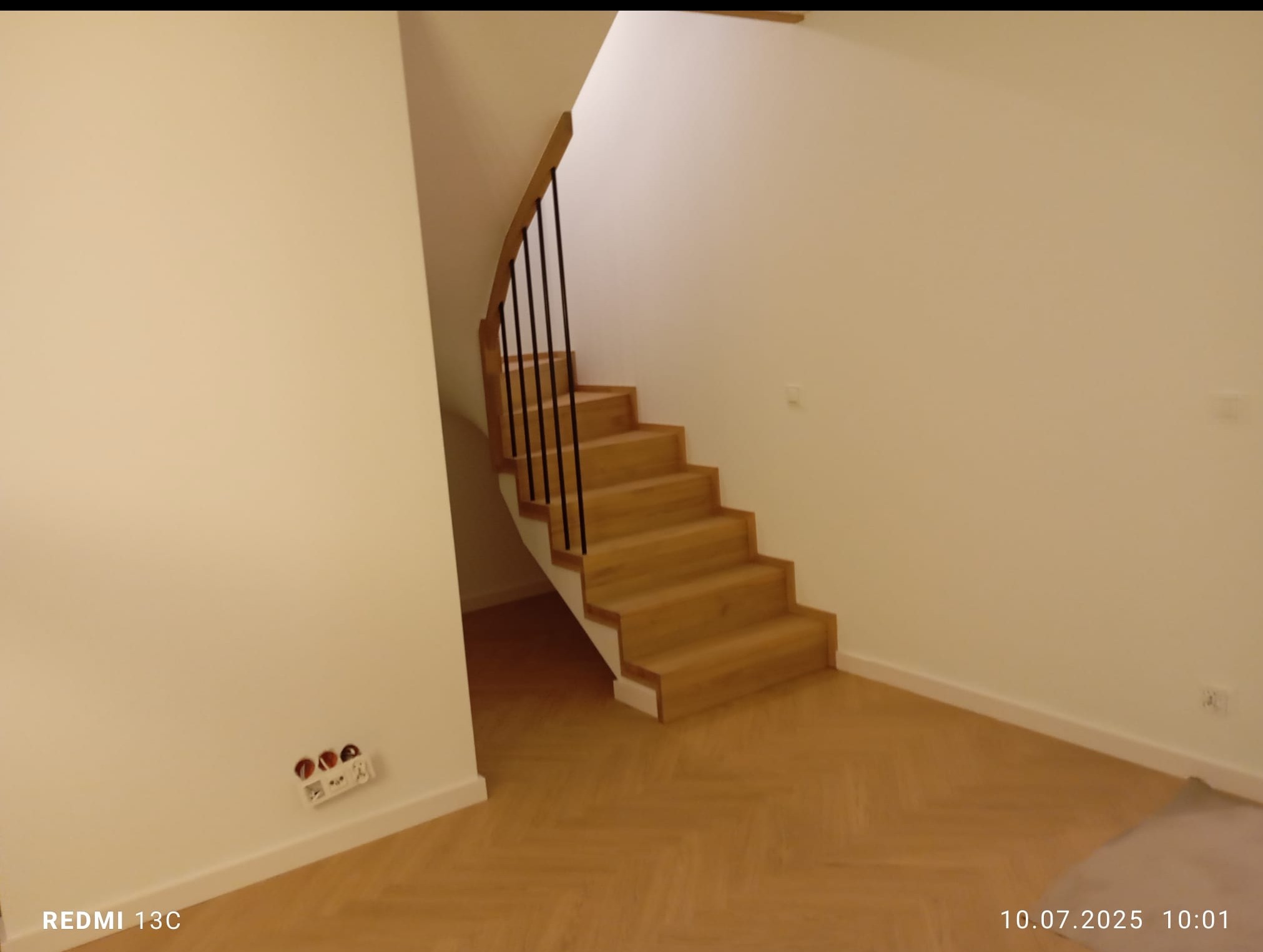 Jasne drewniane schody z czarnymi balustradami w nowym wnętrzu. Parkiet ułożony w jodełkę. Ściany pomalowane na biało. Widoczne gniazdka elektryczne.