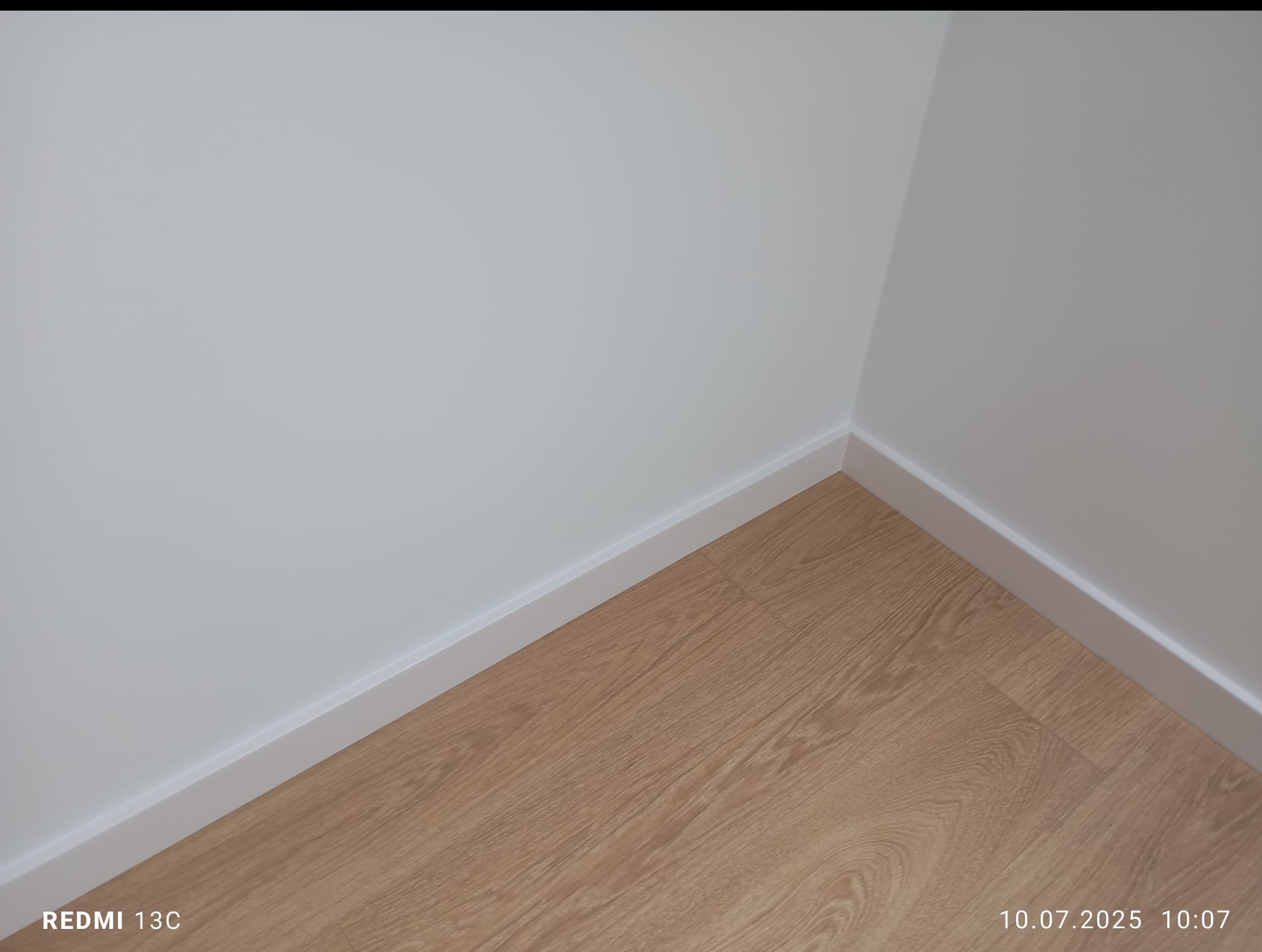 Jasne panele podłogowe w rogu pomieszczenia, białe ściany z przypodłogowymi listwami. Minimalistyczne wykończenie wnętrza w neutralnej kolorystyce.