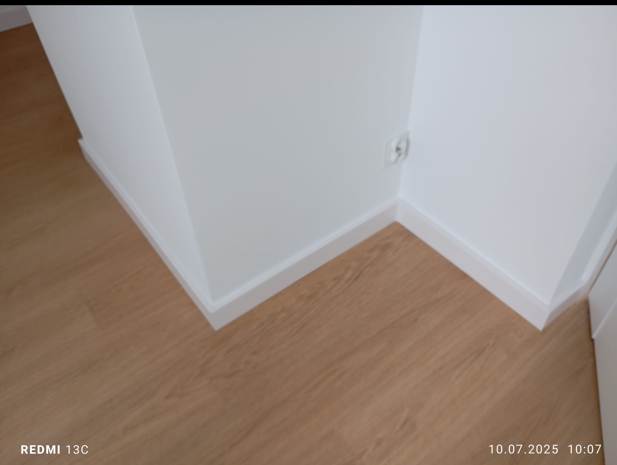 Jasne panele podłogowe i białe listwy przypodłogowe w rogu pomieszczenia. Widoczne gniazdko elektryczne. Minimalistyczny design.