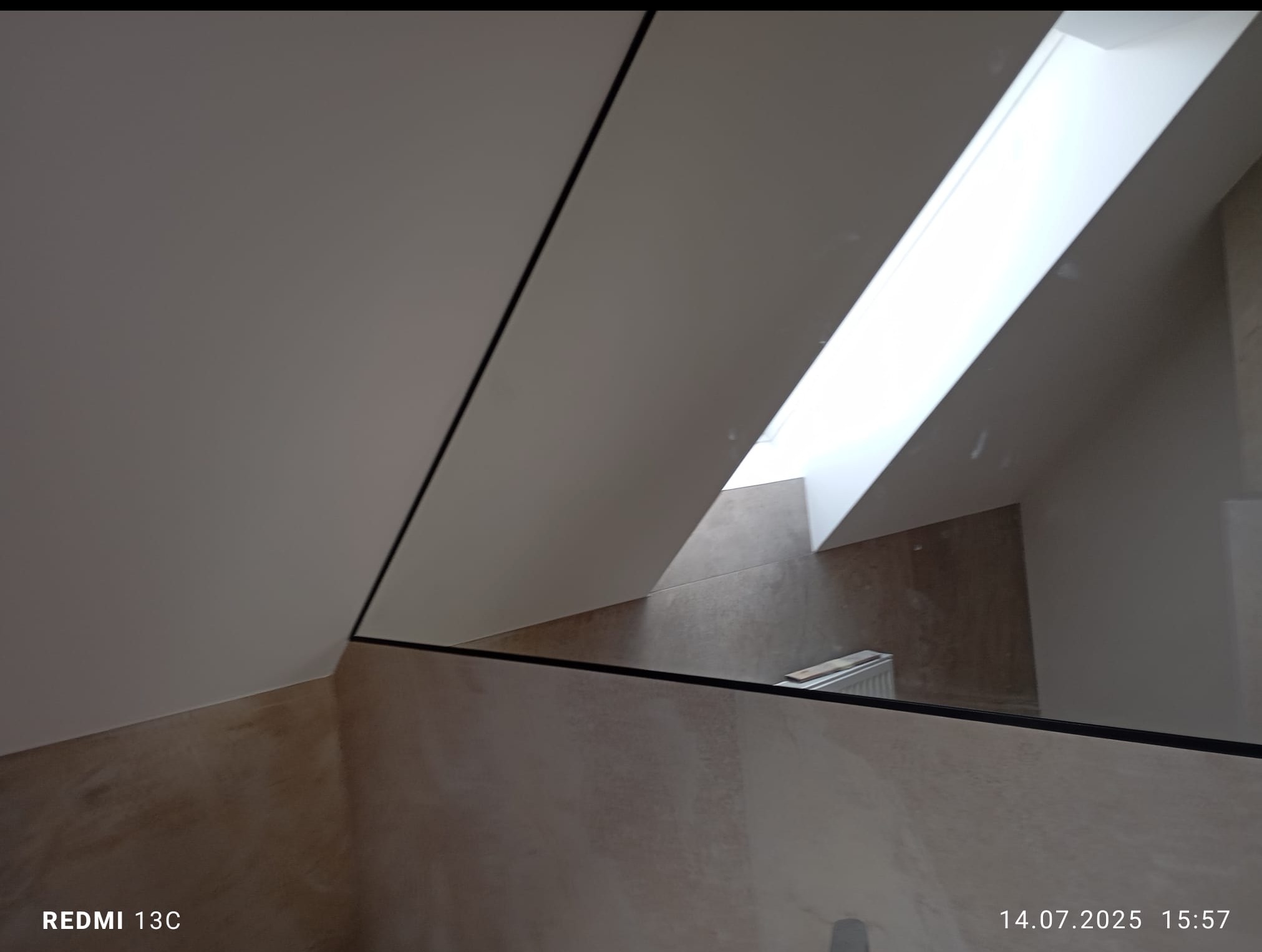 Wykończona łazienka na poddaszu z oknem dachowym, widok przez lustro z czarną ramą. Beżowe płytki na ścianach, biały sufit ze skosami. Minimalistyczny styl.