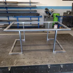 Metalowa konstrukcja stołu w stylu loft na perforowanym stole spawalniczym, z narzędziami i materiałami w tle. Surowy, industrialny charakter warsztatu.