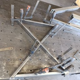 Welddesigner Tomasz Piecyk - Spawanie ramy z profili stalowych na perforowanym stole spawalniczym, z użyciem ścisków stolarskich. Widoczna miarka i profile do spawania.