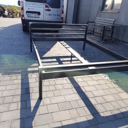 Welddesigner Tomasz Piecyk - Metalowa konstrukcja łóżka na kostce brukowej w słoneczny dzień. W tle van z logo firmy i szary budynek. Widoczne elementy montażowe.