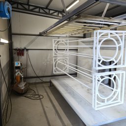 Welddesigner Tomasz Piecyk - Metalowe elementy dekoracyjne, w kwadratowych ramach z okrągłym wzorem w środku, zawieszone w warsztacie, świeżo po malowaniu na biało.