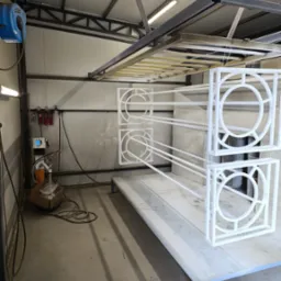 Metalowe elementy dekoracyjne, w kwadratowych ramach z okrągłym wzorem w środku, zawieszone w warsztacie, świeżo po malowaniu na biało.