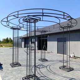 Welddesigner Tomasz Piecyk - Czarna, metalowa konstrukcja altany ogrodowej na szarym bruku, na tle budynku z szarą elewacją i błękitnym niebem w słoneczny dzień. Elementy spawane.