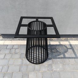 Welddesigner Tomasz Piecyk - Czarna, metalowa konstrukcja przypominająca stolik z ażurowym cylindrem i kwadratową ramą na blacie, stojąca na szarej kostce brukowej.