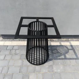 Czarna, metalowa konstrukcja przypominająca stolik z ażurowym cylindrem i kwadratową ramą na blacie, stojąca na szarej kostce brukowej.