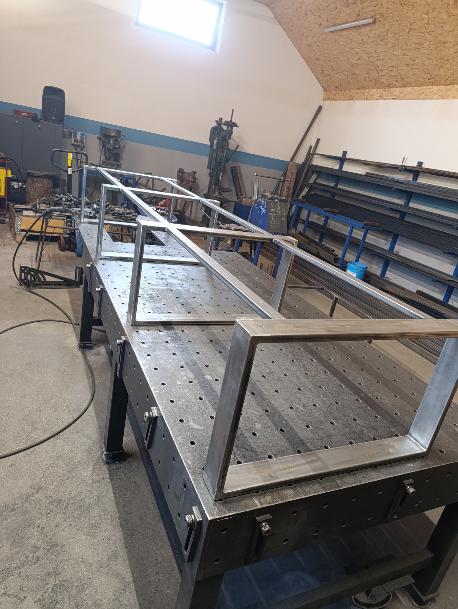 Surowa, metalowa konstrukcja balustrady na perforowanym stole spawalniczym w warsztacie. Widoczne narzędzia i profile metalowe w tle. Górne światło.