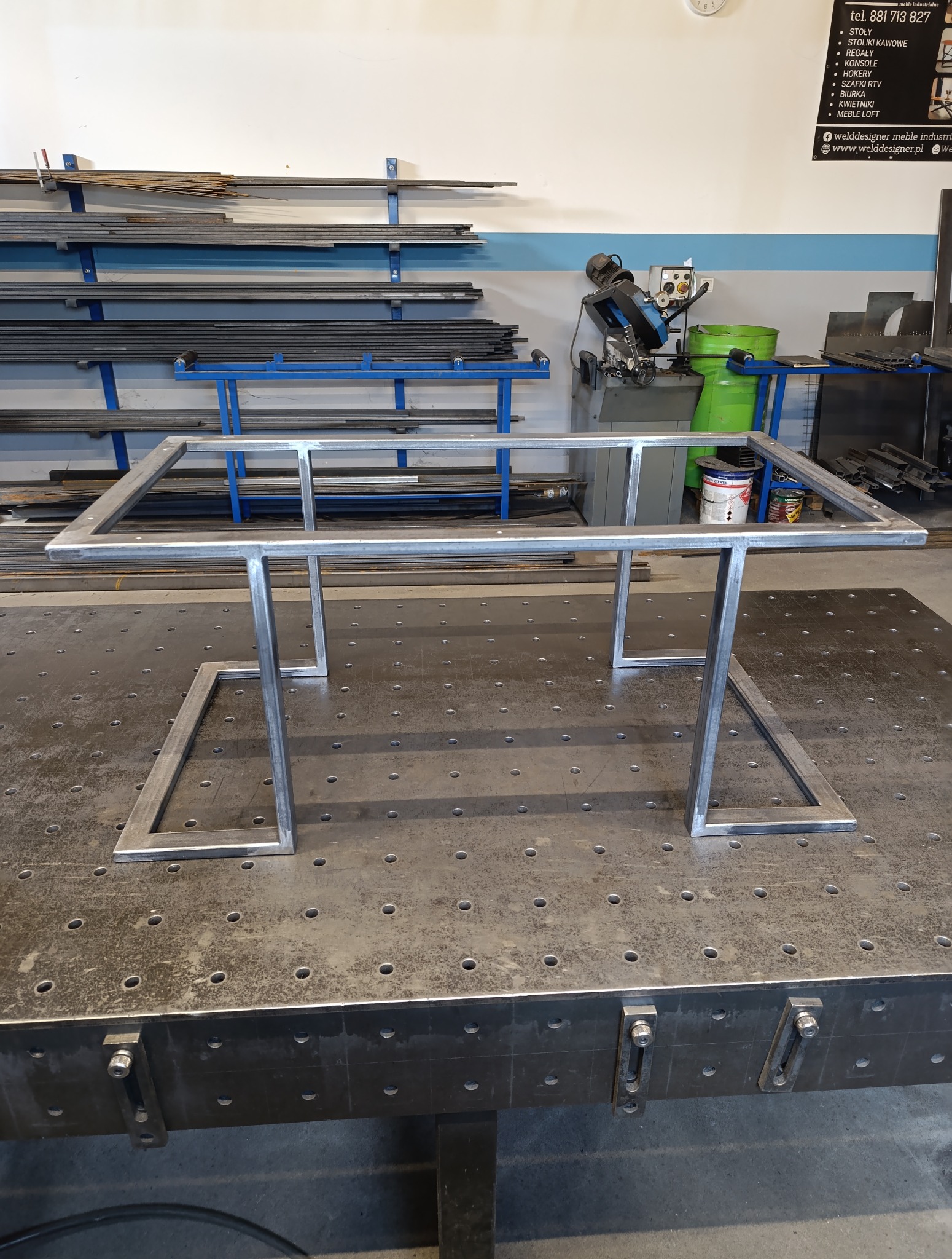 Metalowa konstrukcja stołu w stylu loft na perforowanym stole spawalniczym, z narzędziami i materiałami w tle. Surowy, industrialny charakter warsztatu.