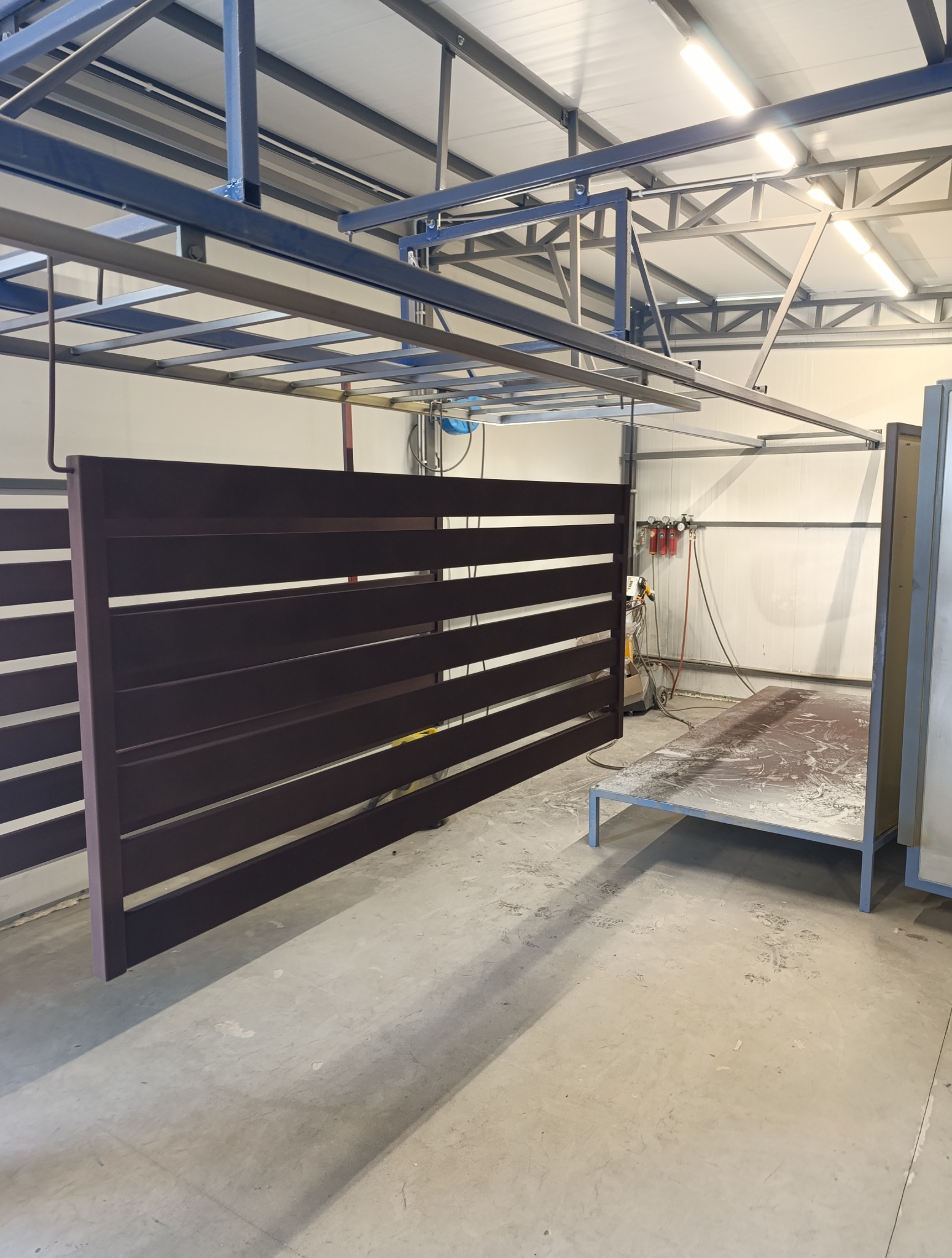 Brązowe, poziome panele ogrodzeniowe zawieszone w hali produkcyjnej, prawdopodobnie po malowaniu. Widoczna konstrukcja sufitowa i stanowisko robocze.