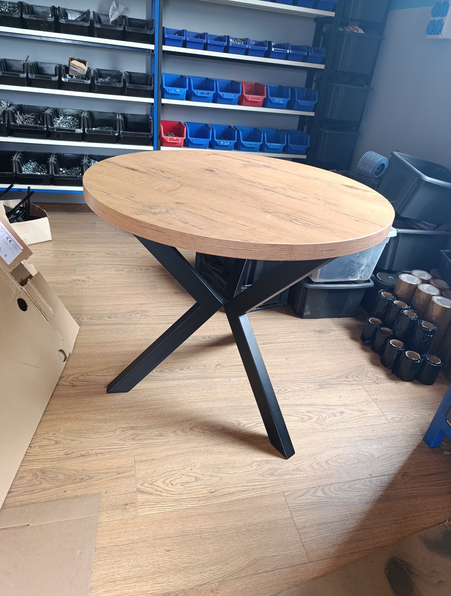 Okrągły stół z drewnianym blatem i czarnymi, metalowymi nogami w kształcie litery X, na tle warsztatu z półkami i pojemnikami na części.