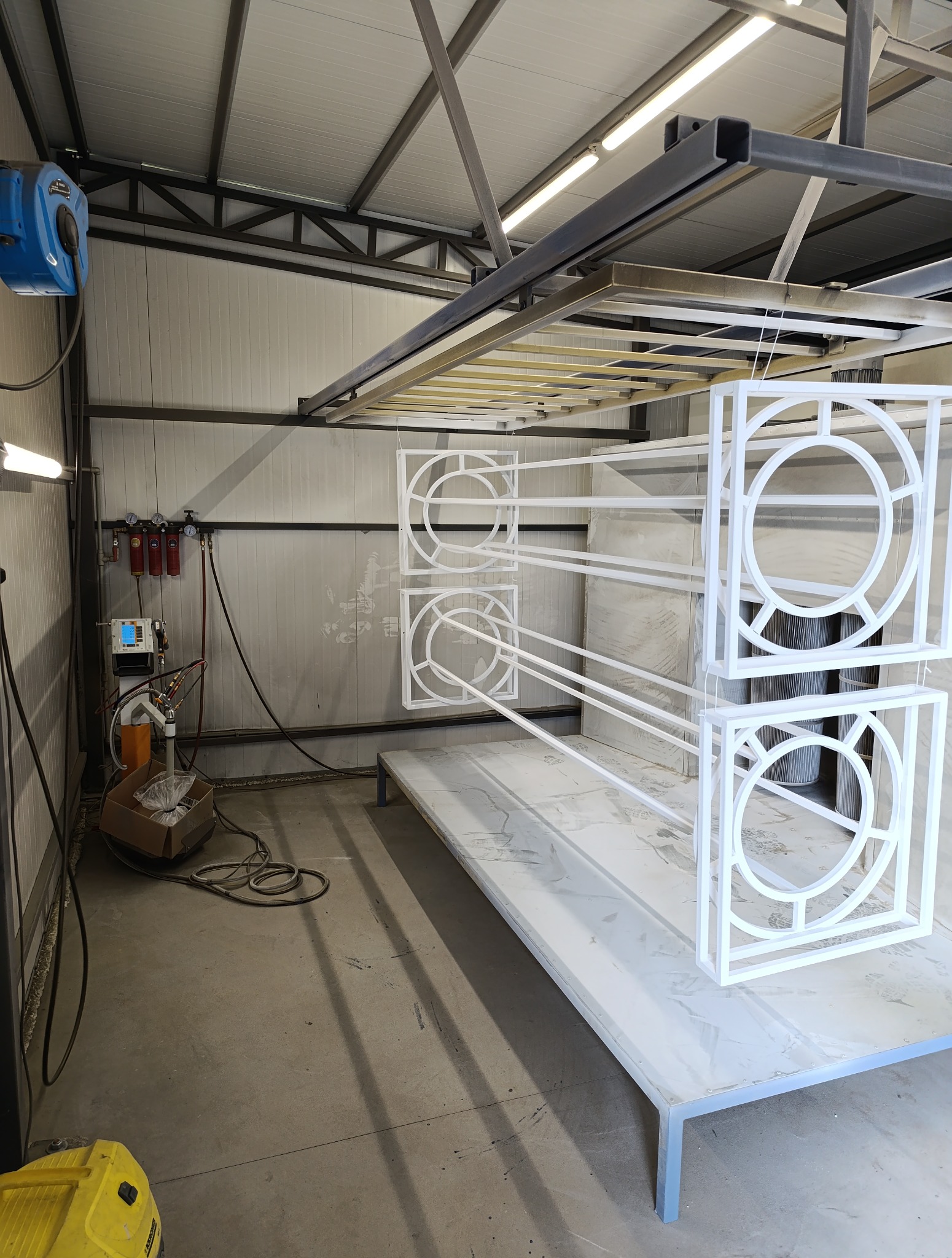 Metalowe elementy dekoracyjne, w kwadratowych ramach z okrągłym wzorem w środku, zawieszone w warsztacie, świeżo po malowaniu na biało.