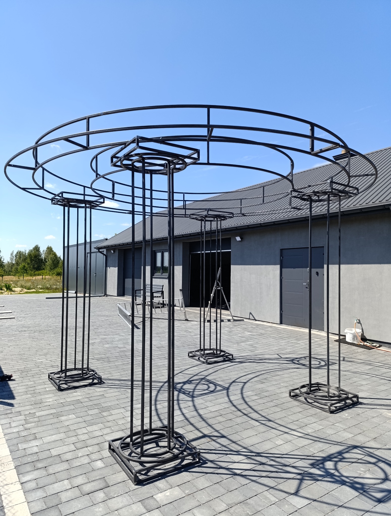 Czarna, metalowa konstrukcja altany ogrodowej na szarym bruku, na tle budynku z szarą elewacją i błękitnym niebem w słoneczny dzień. Elementy spawane.