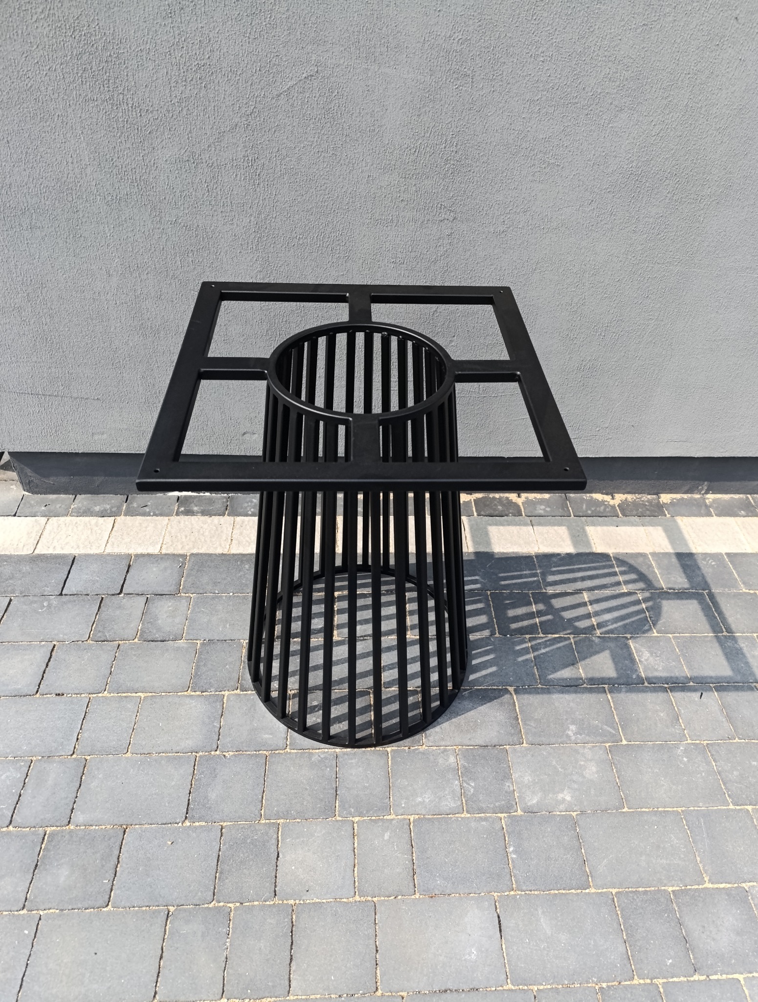 Czarna, metalowa konstrukcja przypominająca stolik z ażurowym cylindrem i kwadratową ramą na blacie, stojąca na szarej kostce brukowej.