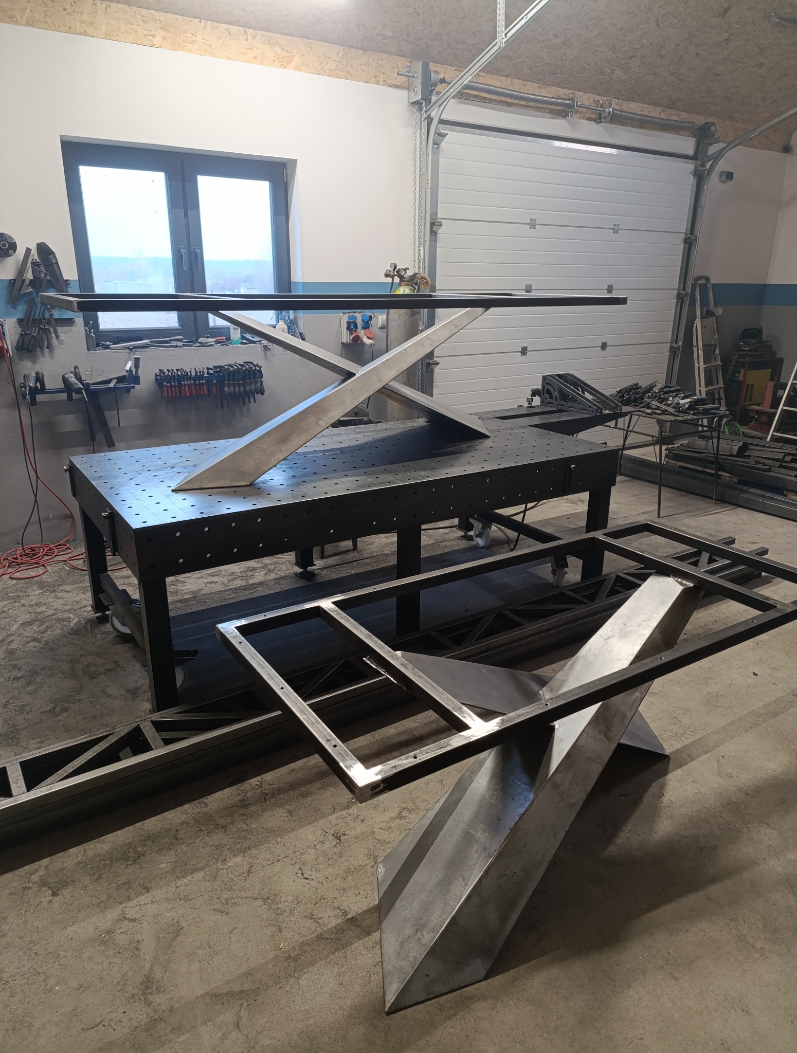 Warsztat z metalowymi elementami konstrukcyjnymi stołów. Dwa blaty i podstawa w trakcie montażu. Widoczne narzędzia i wyposażenie warsztatu.