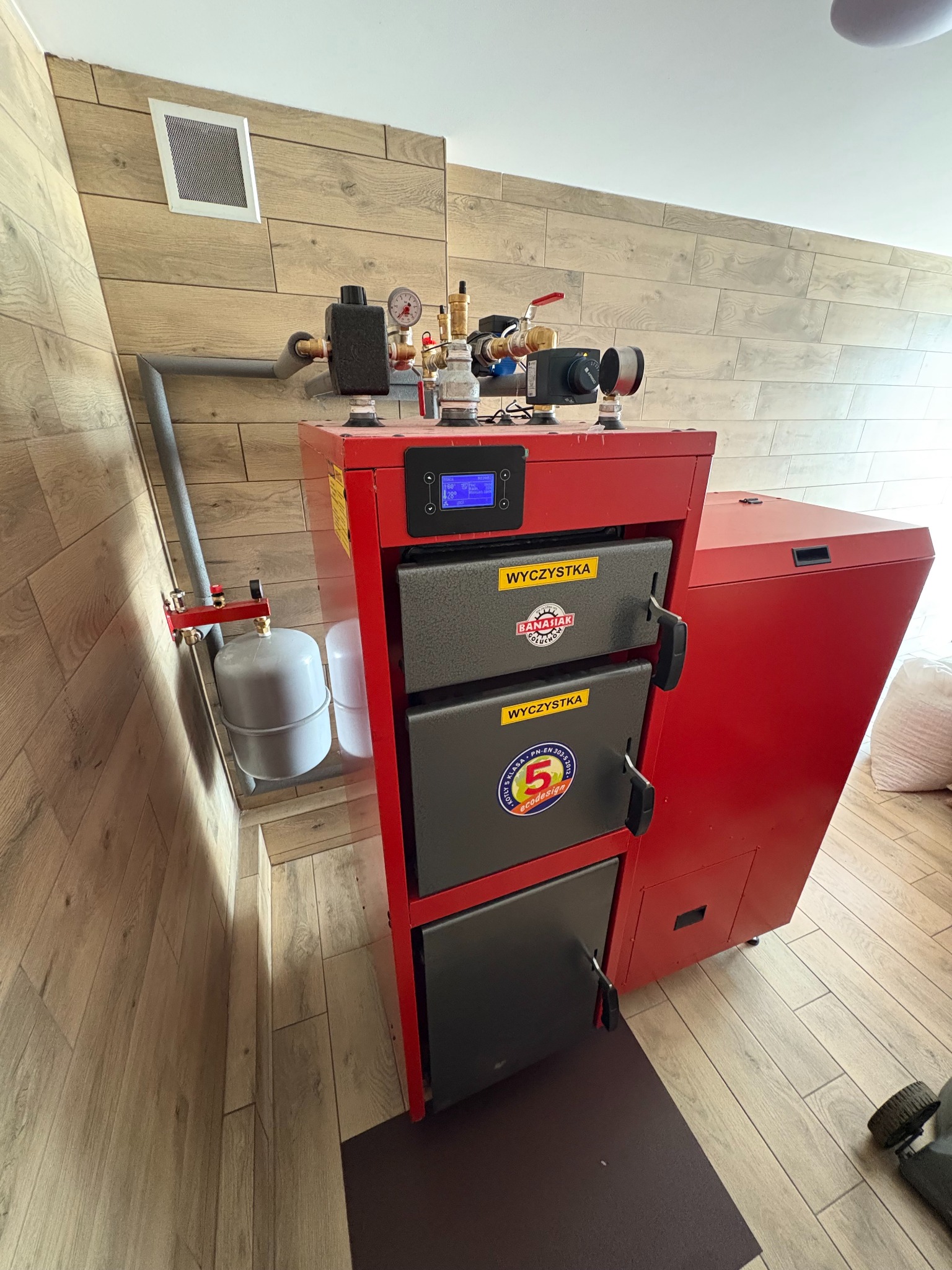 Czerwony piec CO z szarymi drzwiczkami w kotłowni z drewnopodobnymi panelami ściennymi i podłogowymi, widoczny zbiornik wyrównawczy i rury instalacji.