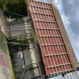 Dach w trakcie krycia: czerwona membrana dachowa z drewnianą łatą, widoczne rusztowania, drabina i fragment ogrodu. Ujęcie z dołu, pochmurne niebo.