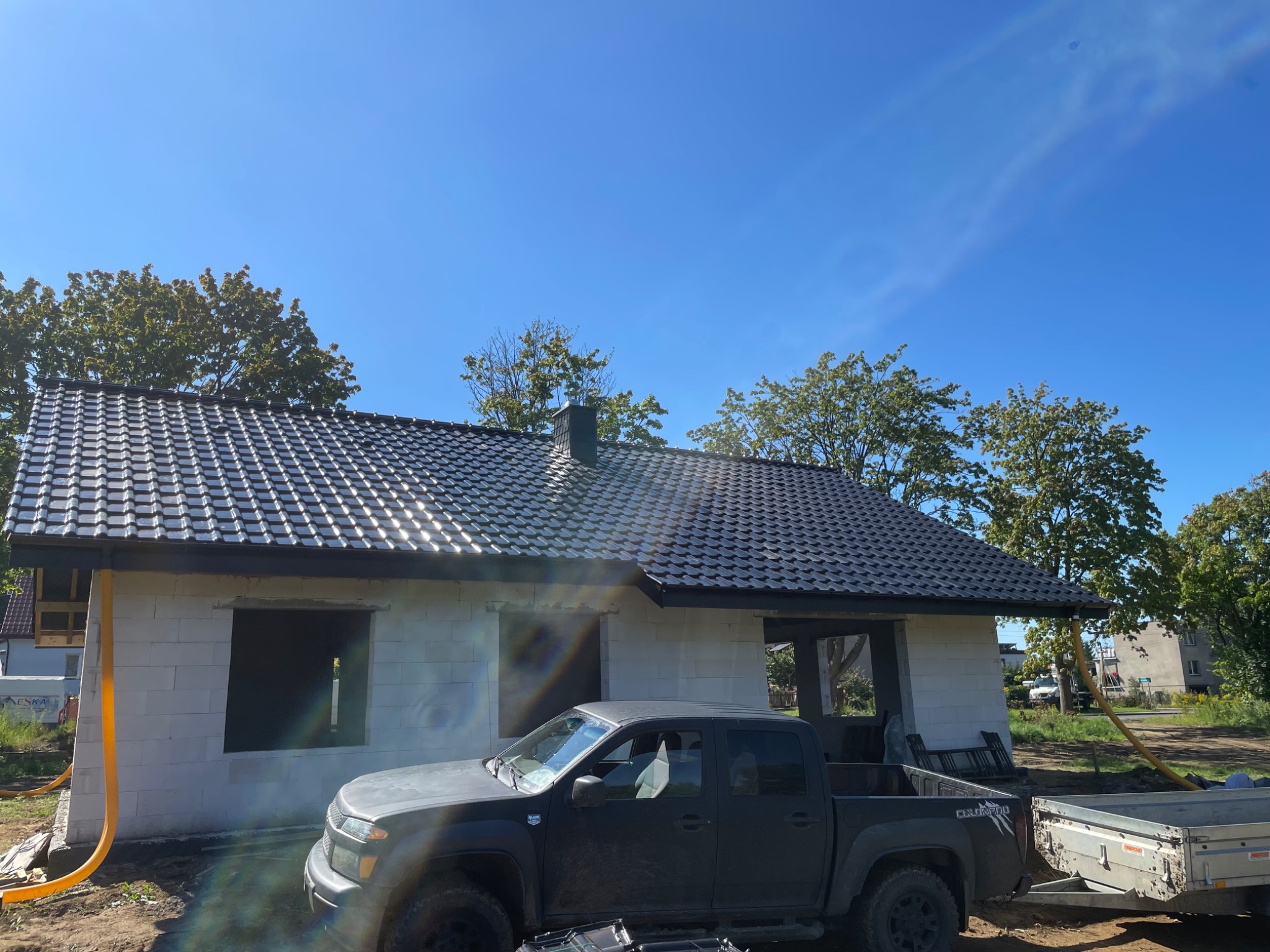 Dom w trakcie budowy z czarnym dachem i ciemnym pickupem na pierwszym planie, błękitne niebo w tle.