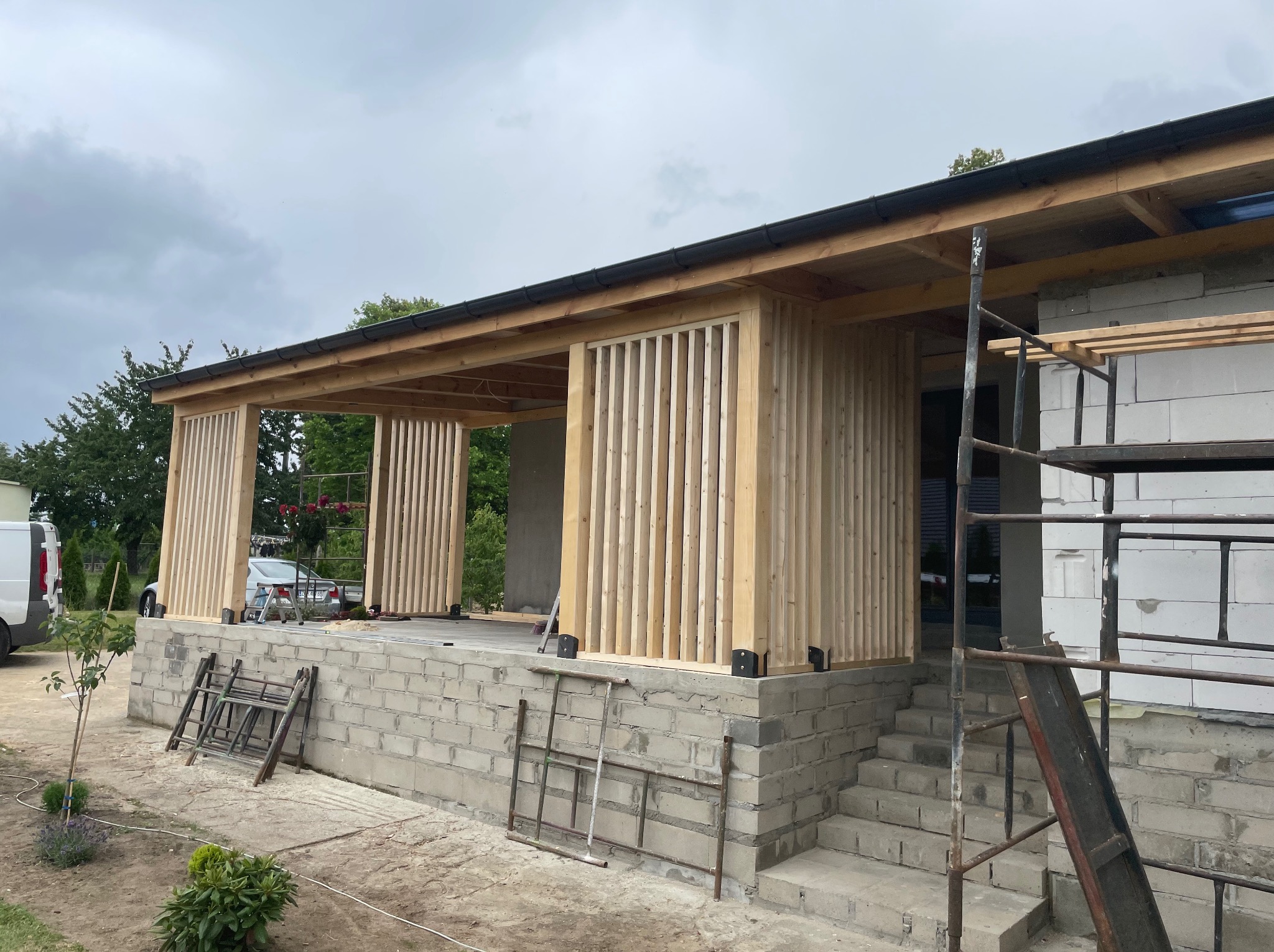 Budowa zadaszonego tarasu z drewnianymi lamelami i schodami z bloczków betonowych. Widoczne rusztowania i materiały budowlane. Ujęcie z dołu, pochmurne niebo.