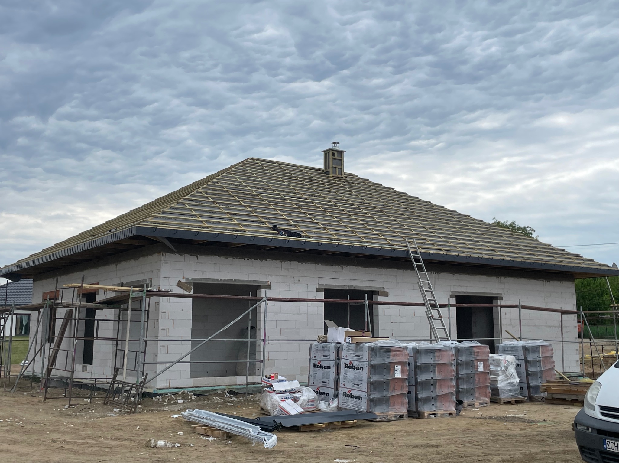 Dom w trakcie budowy z widocznym szkieletem dachu, paletami z dachówkami Roben i rusztowaniem. Pochmurne niebo w tle.