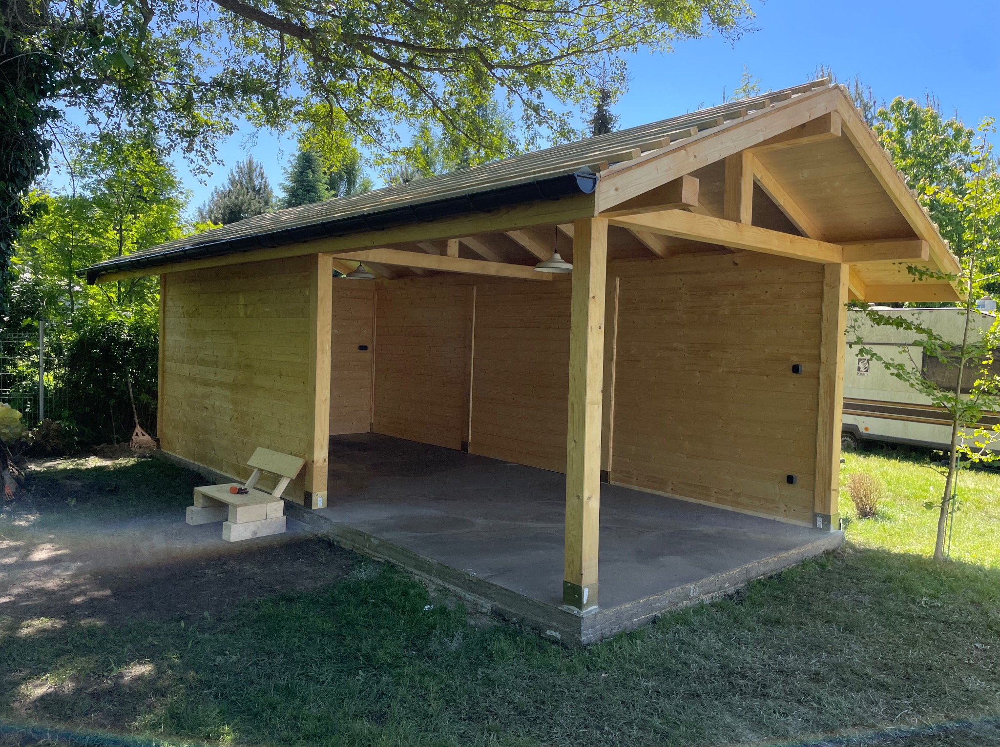 Drewniana konstrukcja z czarną rynną w ogrodzie, częściowo zadaszona. Widoczne elementy szkieletu dachu i podłoże betonowe. W tle zieleń i pojazd kempingowy.
