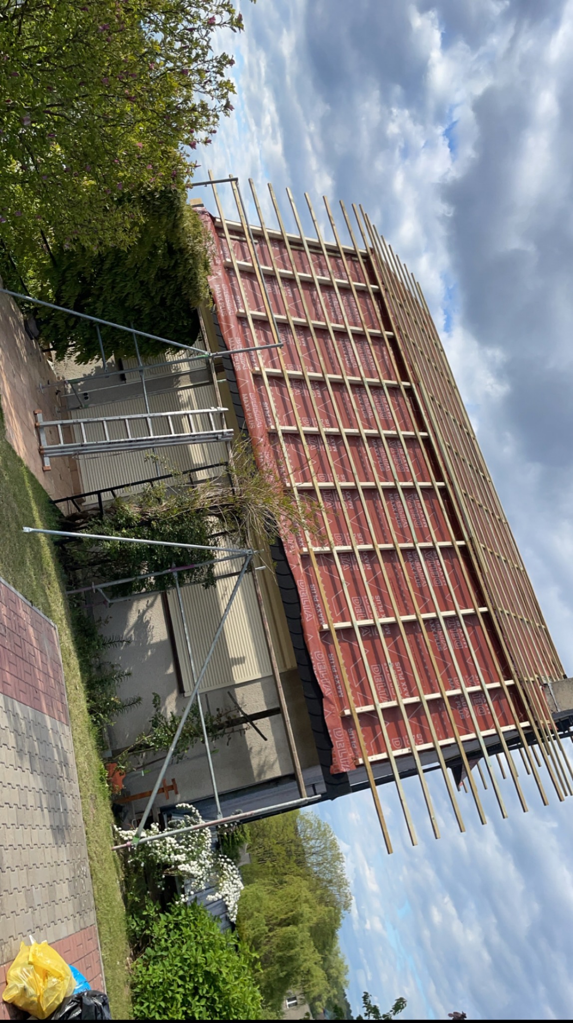 Dach w trakcie krycia: czerwona membrana dachowa z drewnianą łatą, widoczne rusztowania, drabina i fragment ogrodu. Ujęcie z dołu, pochmurne niebo.