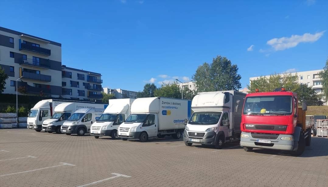 Flota pojazdów transportowych, w tym ciężarówka DAF, zaparkowana na parkingu z kostką. Na zabudowach aut reklama Castorama. Budynek mieszkalny w tle.