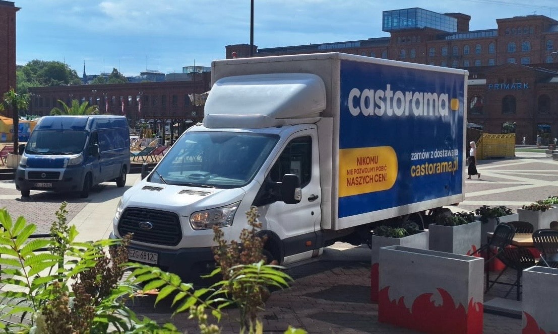 Biały bus dostawczy Castorama z niebieską reklamą na tle miejskiego placu z ceglaną architekturą. Drugi, mniejszy bus w tle. Słoneczny dzień.